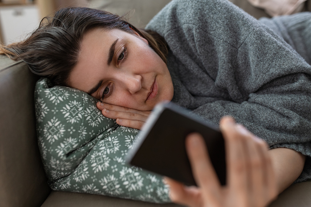 a woman lies on the couch doomscrolling on her phone.